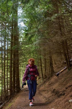 Uzun çam ağaçları ve doğal çalılıklarla çevrili bir ormanda tek başına yürüyen bir kadın.