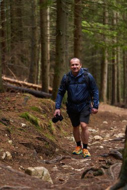 Sırt çantalı ve kameralı bir adam engebeli arazide bir orman patikasına tırmanıyor, odaklanmış ve açık hava fotoğrafçılığı için donanımlı.
