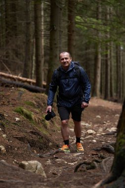Sırt çantalı ve kameralı bir adam engebeli arazide bir orman patikasına tırmanıyor, odaklanmış ve açık hava fotoğrafçılığı için donanımlı.