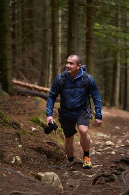 Sırt çantalı ve kameralı bir adam engebeli arazide bir orman patikasına tırmanıyor, odaklanmış ve açık hava fotoğrafçılığı için donanımlı.