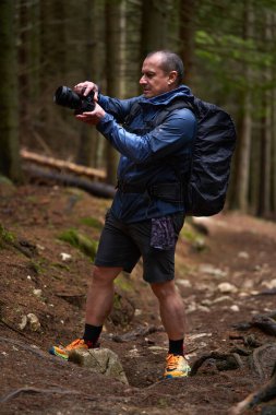 Elinde kamera olan bir adam engebeli bir patikada dikilirken ormanı fotoğraflıyor.