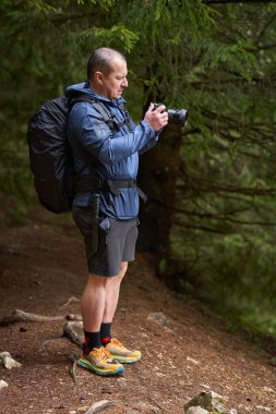 Elinde kamera olan bir adam engebeli bir patikada dikilirken ormanı fotoğraflıyor.
