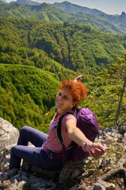 Gülümseyen kızıl saçlı kadın, orman manzarasının üzerindeki kayalık bir uçurumun kenarında otururken geniş kollarıyla dağ manzarasının tadını çıkarıyor.