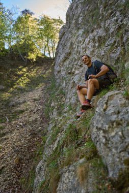 Sırt çantalı bir adam bir dağ yürüyüşü sırasında dik bir orman yamacı boyunca kayalık bir çıkıntıda otururken gülümsüyor.