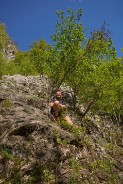 Sırt çantalı bir adam bir dağ yürüyüşü sırasında dik bir orman yamacı boyunca kayalık bir çıkıntıda otururken gülümsüyor.