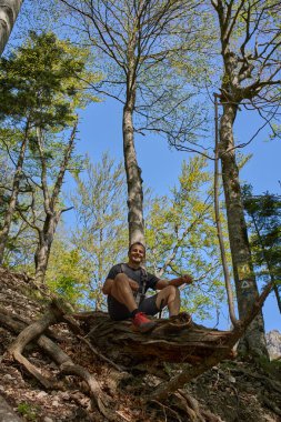 Sırt çantalı gülümseyen bir adam güneşli bir bahar yürüyüşü sırasında geniş bir ağaç gövdesinin üzerinde, bir orman yamacında korunmasız kökler arasında oturuyor.