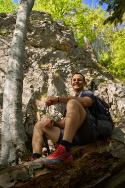 Sırt çantalı gülümseyen bir adam güneşli bir bahar yürüyüşü sırasında geniş bir ağaç gövdesinin üzerinde, bir orman yamacında korunmasız kökler arasında oturuyor.