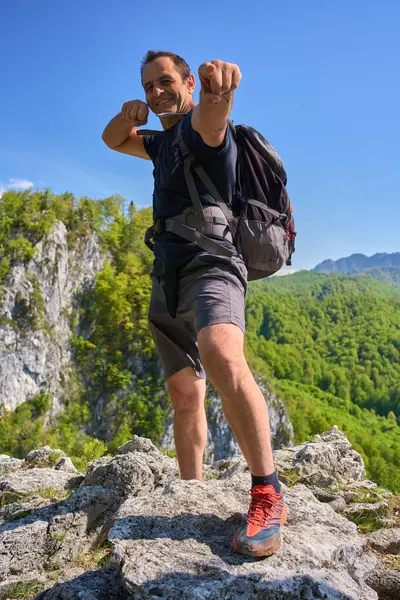 Sırt çantalı gülümseyen adam, açık mavi gökyüzünün altındaki yeşil ormanla çevrili kayalık bir dağ manzarasında kamerayı işaret ediyor.