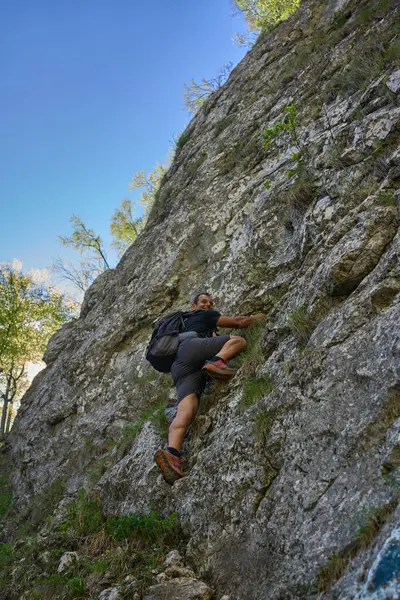 Sırt çantalı bir erkek dağcı, ormanlık bir bölgede güneşli bir günde, dik bir kayalık yamaçta doğal tutarak yükseliyor.