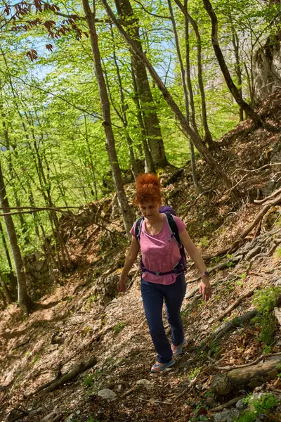 Sırt çantalı bir kadın, yeşil ağaçlarla çevrili bahar güneşinde, kökleriyle kaplı bir orman patikasında yokuş yukarı yürüyor.