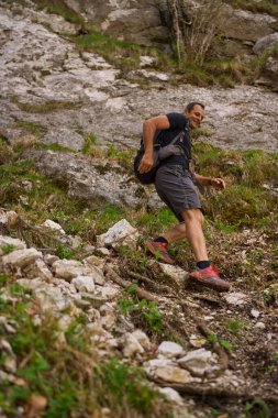Dik bir yamaçtan inen bir adam yürüyüş kıyafetleri ve kırmızı patika koşu ayakkabıları giyiyor.
