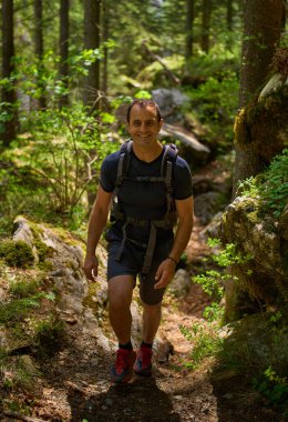 Yosun kaplı kayalar ve bahar bitkileriyle çevrili bir orman yolunda yokuş yukarı yürüyen bir adam.