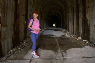 Sırt çantalı, ıslak beton zemin ve kayalık duvarları olan kavisli bir tünelin içinde gülümseyen bir kadın.