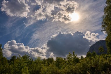 Kümülüs ve sirrus bulutları bir bahar ormanının üzerinde. Güneş kısmen gizlenmiş ve arka planda kayalık bir dağ yamacı var.