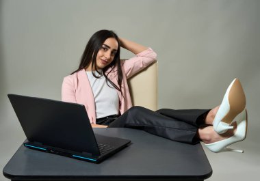 Kendine güveni tam bir iş kadını, bacakları masanın üzerinde ve rahat duruşu dizüstü bilgisayarın arkasında, gri arka planda tek başına.