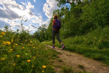 Çamur patikasında tek başına yürüyüş yapan bir kadın dramatik bir gökyüzü altında yeşil ve kır çiçekleriyle çevrili, alçak bir açıdan vurulmuş.