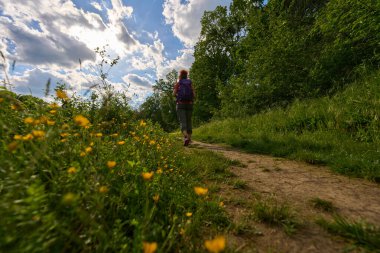 Çamur patikasında tek başına yürüyüş yapan bir kadın dramatik bir gökyüzü altında yeşil ve kır çiçekleriyle çevrili, alçak bir açıdan vurulmuş.