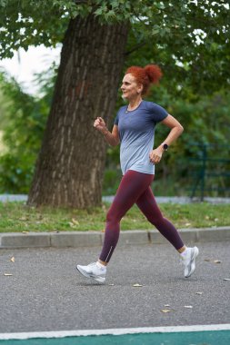 Açık hava antrenmanı sırasında ağaçlarla çevrili, kaldırımlı yolda koşan olgun bir kadın.