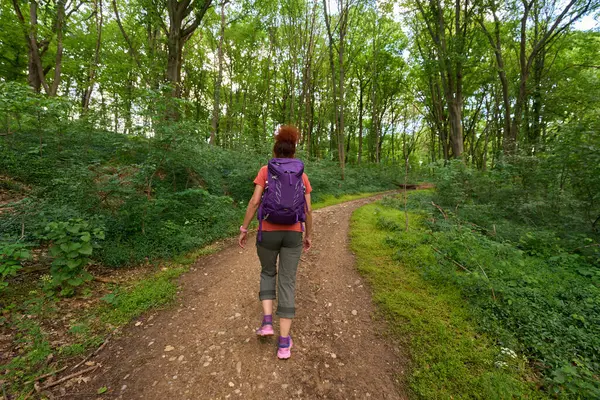 Sırt çantalı bir kadın bir bahar günü orman yolunda tek başına yürüyüş yapıyor.