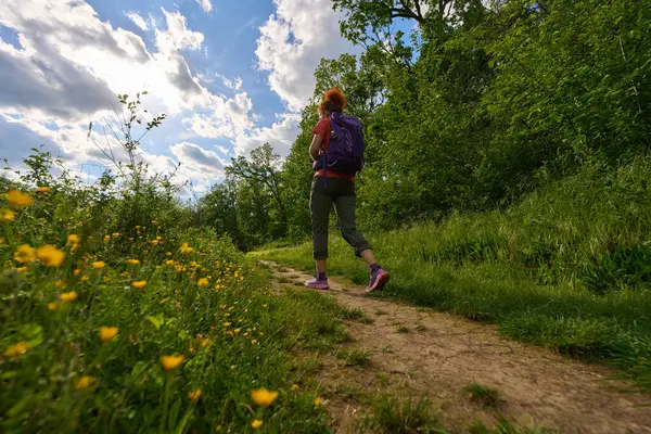 Çamur patikasında tek başına yürüyüş yapan bir kadın dramatik bir gökyüzü altında yeşil ve kır çiçekleriyle çevrili, alçak bir açıdan vurulmuş.