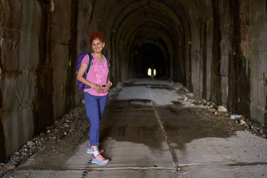 Sırt çantalı kadın, yürüyüş yolunda taş tünelin içinde dikilirken güvenle gülümsüyor.