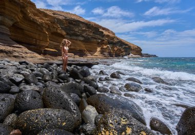 Çiçekli bikinili genç bir kadın Tenerife 'deki Playa de Montana Amarilla' da volkanik kayalarda yalınayak duruyor. 