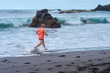 Çıplak ayakla koşan turuncu tişörtlü bir adam ıslak volkanik plajda okyanus dalgalarıyla sıçrıyor.