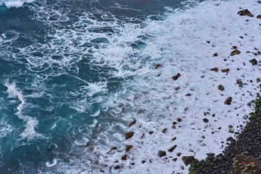 Tenerife 'deki El Bollullo plajının yanındaki kayalık volkanik kıyı şeridinin hava manzarası yosun kaplı kayaların üzerine köpüklü dalgalar düşüyor.