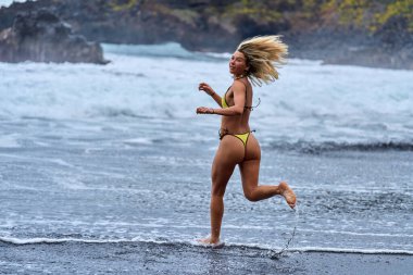 Sarı bikinili gülümseyen kadın Tenerife 'deki volkanik plajda sığ dalgalarda neşeyle koşuyor.