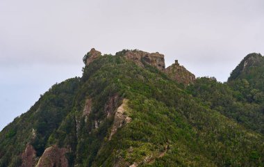 Kayalık zirvesiyle yemyeşil tepe ve bulutlu bir gökyüzünün altında ormanlı yamaçlar.