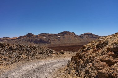 Açık mavi gökyüzünün altında Teide Ulusal Parkı 'nın içindeki çorak bir patikadan görülen katmanlı volkanik tepe.