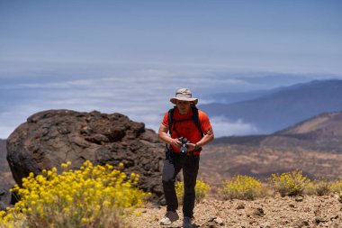 Kayalık volkanik arazide elinde kamerayla yürüyen ve arka planda dağ manzaralı bir adam.