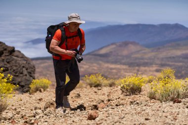 Kayalık volkanik arazide elinde kamerayla yürüyen ve arka planda dağ manzaralı bir adam.