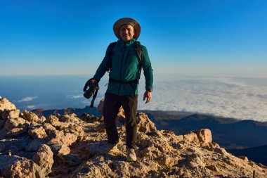 Pico del Teide zirvesinde duran sırt çantalı ve kameralı adam gün batımında volkanik manzarayı ve bulutun tersini yakalıyor.