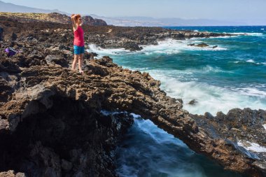 Atlantik kıyısına bakan aşınmış lav kemerinde duran engebeli dalgaları ve volkanik arazisi olan bir kadın.