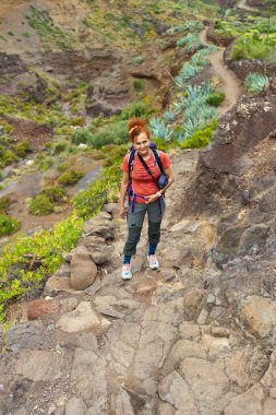 Sırt çantalı ve kameralı bir kadın Tenerife 'nin Anaga dağlarındaki kayalık yolda duruyor. Kuru subtropikal araziyle çevrili.