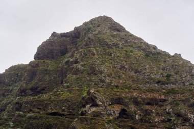 Tenerife 'de bulutlu bir gökyüzünün altında aşınmış kayalık oluşumları olan yeşil volkanik tepeler.