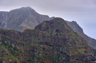 Anaga 'da volkanik dağ sıraları. Yerleşik bitkiler ve bulutlu bir gökyüzünün altındaki aşınmış kayalıklar.