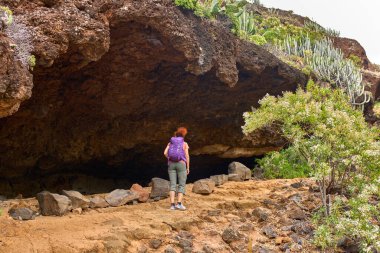 Mor sırt çantalı bir kadın Tenerife 'deki kayalık yolda volkanik mağaraya yaklaşıyor.