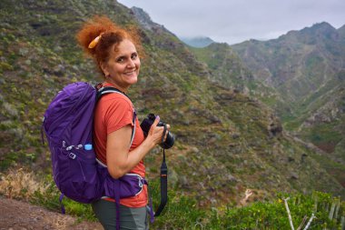 Anaga yolunda kamerayla yürüyüş yapan bir kadın dağ manzarasının fotoğrafını çekmek için duruyor.