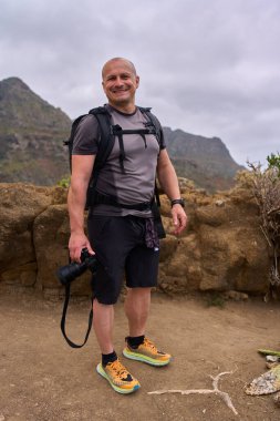 Tenerife 'de yürüyüş yolunda gülümseyen bir adam elinde bir kamera ve arkasında volkanik dağlar olan bir sırt çantası tutuyor.