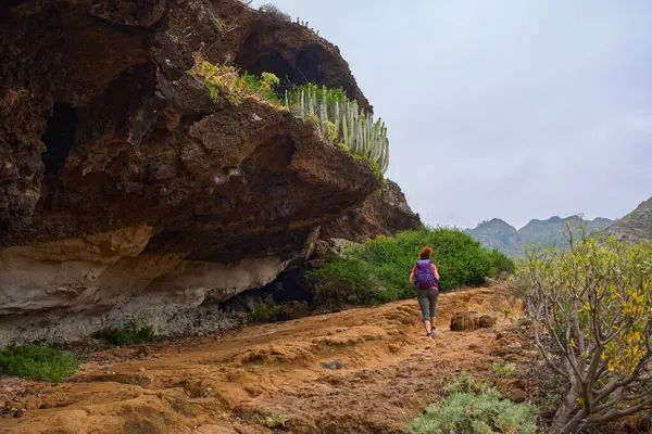 Mor sırt çantalı kadın yürüyüşçü Tenerife 'deki mağaranın yakınındaki kayalık volkanik patikayı keşfediyor.