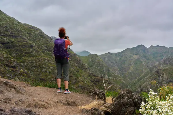 Anaga yolunda kamerayla yürüyüş yapan bir kadın dağ manzarasının fotoğrafını çekmek için duruyor.