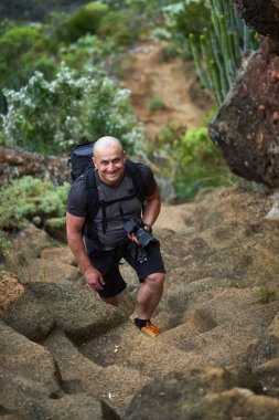 Gülümseyen erkek yürüyüşçü Tenerife 'de kamera tutarken volkanik patikaya tırmanıyor