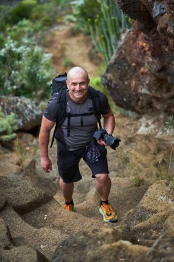 Gülümseyen erkek yürüyüşçü Tenerife 'de kamera tutarken volkanik patikaya tırmanıyor