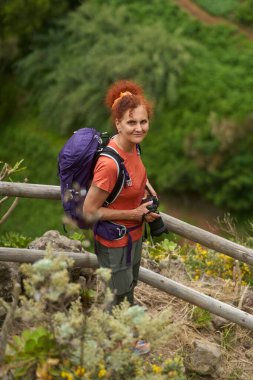Kamera ve mor sırt çantalı kızıl saçlı bayan yürüyüşçü Tenerife 'de yolda gülümsüyor.