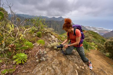 Anaga 'daki dağ yolunda diz çöken kadın fotoğrafçı arka planda okyanusla birlikte yerli bitki örtüsünü yakına çekiyor.