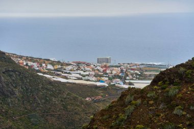 Punta del Hidalgo kasabasının uzak manzarası ve volkanik bitki örtüsüyle yüksek bir yamaçtan görünen kıyı şeridi.