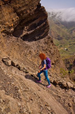 Kadın, Tenerife 'deki Teno' nun güneşli dağlarında aktif bir yürüyüş sırasında kayalık bir patikaya tırmanıyor.