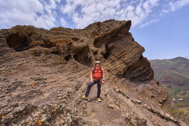 Gülümseyen yürüyüşçü aşınmış volkanik uçurumun ve Tenerife patikasındaki mağaraların önünde poz veriyor.